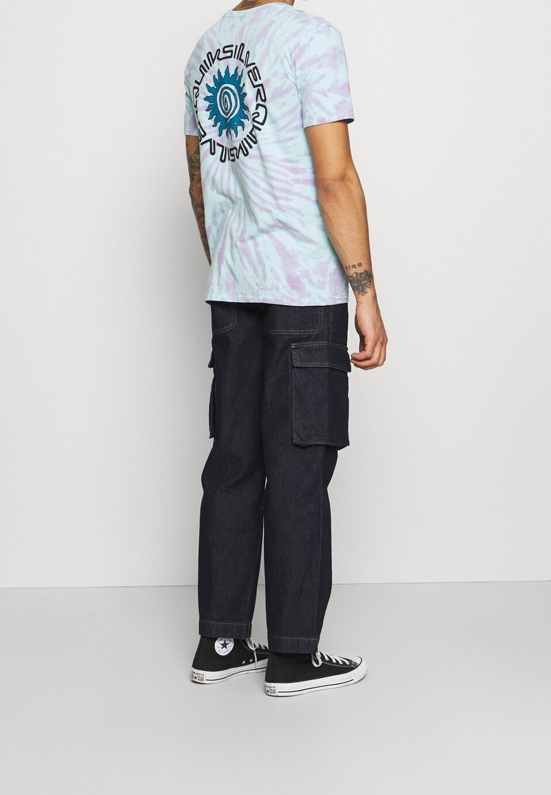 Ljus lila tie-dye bomullströja med blå grafik på baksidan. Mörka denim cargobyxor med stora sidofickor. Svarta högsneakers.