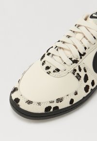 Fehér sneaker fekete foltos mintával, bőrből és texturált anyagból készült. Fekete gumi talp, azonos színű részletekkel. Lapos cipőfűzők.