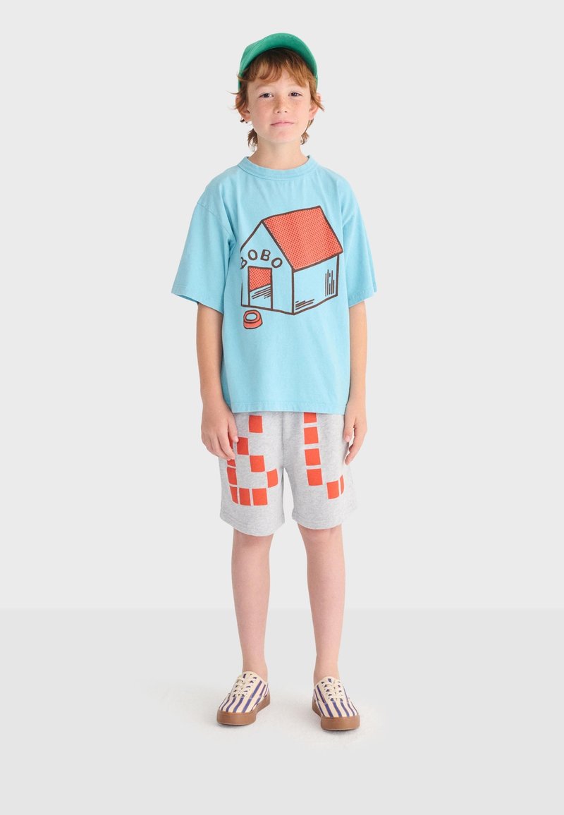 Jeune garçon portant une casquette verte, un t-shirt bleu clair avec un motif de niche, un short gris à carreaux orange et des chaussures slip-on rayées.