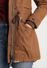 Personne portant une veste marron avec la main dans la poche avant, fermeture éclair visible, boutons pression et cordon de serrage, assortie à un jean bleu.