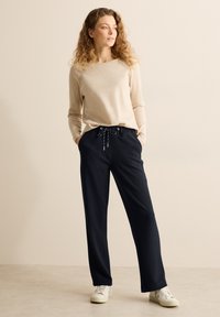 Haut à manches longues beige clair associé à un pantalon noir avec cordon de serrage ; le pantalon est ample avec des poches, et des baskets blanches complètent le look.