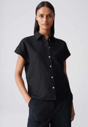 Vrouw met steil donker haar draagt een zwart overhemd met korte mouwen en een zwarte broek tegen een effen lichtgrijze achtergrond.