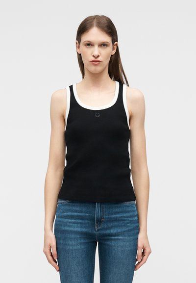 Γυναίκα με ίσια καστανή τρίχα που φοράει μαύρο αμάνικο πάνω μέρος με λευκό περίγραμμα και μπλε τζιν, στέκεται μπροστά σε απλό φωτεινό φόντο.