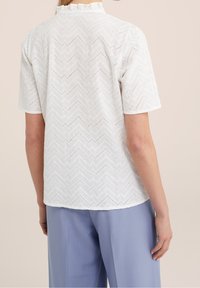 Vit kortärmad blus med ett zickzackmönster, texturerat tyg och en volangkrage. Bärs med ljusblå byxor.