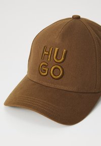 Brown cap made of cotton with a structured design. Features raised, gold embroidery spelling "HU GO" on the front. Curved brim.