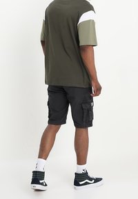 Camisa de manga corta color verde oliva con detalles en blanco y gris, combinada con pantalones cortos cargo negros y zapatillas altas marinas con suelas blancas.