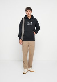 Μαύρη φούτερ με κουκούλα με το γράμμα "VISION" σε λευκή εκτύπωση, συνδυασμένη με μπεζ cargo παντελόνια. Το μοντέλο φοράει μπεζ αθλητικά και μια γκρι τσάντα ώμου.