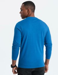 Man i en ljusblå långärmad skjorta, svart klocka på vänster handled, står med ryggen mot kameran och huvudet lätt vridet åt vänster.