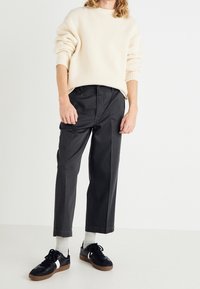 Un pull en tricot beige clair à la coupe ample, associé à un pantalon gris foncé à jambe fuselée. Des baskets noires avec une touche blanche et des chaussettes grises complètent le look.