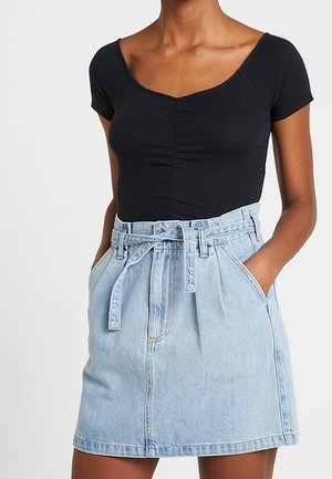 Kvinde iført en sort kortærmet top med rynket midterparti og en lyseblå denimnederdel med bælte og hænder i lommerne.