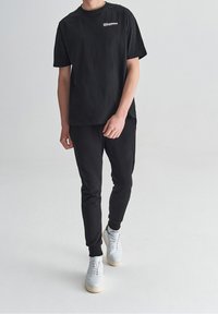 Hombre vistiendo una camiseta negra con el logo "Modern", pantalones de chándal negros y zapatillas blancas, de pie contra un fondo claro y liso.