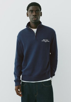 Jeune homme portant un pull bleu marine à fermeture éclair au quart avec le texte "New York Urban Citizen 1991" debout devant un fond clair uni.