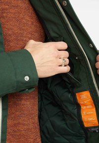 Groene jas met een gewatteerde binnenkant, zilveren rits en oranje label; gedragen over een gemêleerde oranje trui; hand die in de jas reikt.