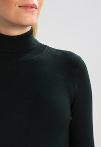 Ciemnozielony sweter z golfem wykonany z materiału ze splocie prążkowanym. Posiada dopasowany krój z wysokim kołnierzem i długimi rękawami.