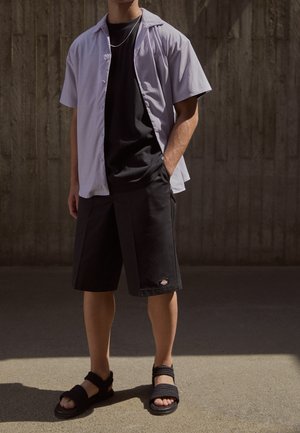 Camisa de botones de mangas cortas en morado claro sobre una camiseta negra, combinada con pantalones cortos negros y sandalias negras con textura.