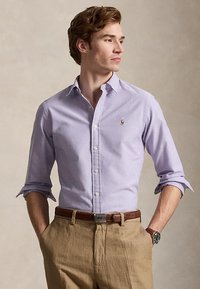 Férfi világoslila gombos ingben, felhajtott ujjakkal, bézs nadrágban, barna övvel és órával, zsebébe tett kézzel áll.