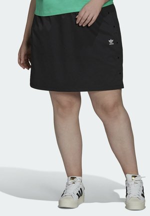Person iført en sort Adidas nederdel med knapper i siden, hvide Adidas sneakers med sorte striber og en grøn skjorte, stående.