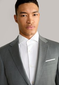Graue Anzugjacke mit Revers und einem weißen Einstecktuch. Darunter ein weißes Hemd mit Button-down-Kragen und sichtbaren Knöpfen.