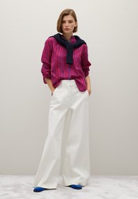 Donna che indossa una camicia a righe verticali rosse e blu, un maglione blu scuro drappeggiato sulle spalle, pantaloni bianchi a gamba larga e scarpe blu, in piedi con le mani nelle tasche.