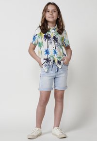 Camisa de manga corta abotonada con estampado colorido de palmeras sobre un fondo crema, combinada con pantalones cortos de denim azul claro y zapatillas blancas.