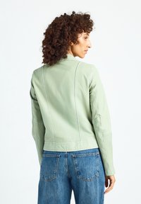 Zielona skórzana kurtka o dopasowanym fasonie, z kontrastującymi białymi przeszyciami i stójką, zestawiona z niebieskimi dżinsami.