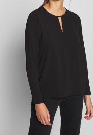 Mujer con una blusa negra de manga larga con una pequeña abertura en forma de ojo de cerradura en la parte delantera y pantalones negros, de pie frente a un fondo liso.