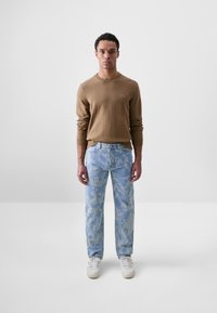 Suéter de punto marrón claro con cuello redondo, combinado con jeans de mezclilla azul claro que presentan un sutil patrón de camuflaje y zapatillas blancas.
