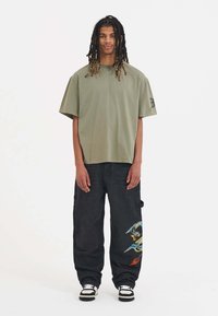 Olive green oversized t-shirt with short sleeves, paired with black cargo pants featuring a colorful dragon embroidery and white sneakers.