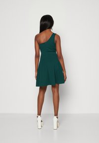 Grön, enaxlad klänning gjord av mjukt tyg med figursydd överdel och utsvängd kjol; parat med vita högklackade skor.