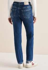 Jeans in denim blu scuro con un design a gamba dritta, dotati di due tasche posteriori e un'etichetta del marchio in vita. Indossati con sneakers bianche.