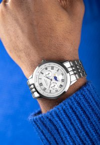Silver wristwatch with a textured silver dial, Roman numeral markers, date and moon phase complications, and a polished metal bracelet.