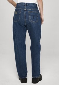 Vaqueros de mezclilla azul oscuro de talle alto con pierna recta. Dos bolsillos traseros con detalles de costura. Botas negras gruesas visibles en la parte inferior.