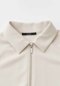 Giacca beige con zip, colletto appuntito e etichetta interna nera con la scritta "someday."