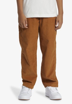 Barn iført brune kordbukser, hvide sneakers og en hvid skjorte, stående med hænderne afslappet ved siden på en hvid baggrund.