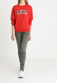 Rotes Sweatshirt mit "RAW" Grafik und gemustertem Schriftzug, kombiniert mit olivgrünen Slim-Fit-Hose und weißen Sneakers.