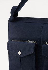 Sac messager en tissu bleu marine avec deux poches zippées à l'avant, fermeture à bouton-pression et surface texturée avec des coutures robustes.