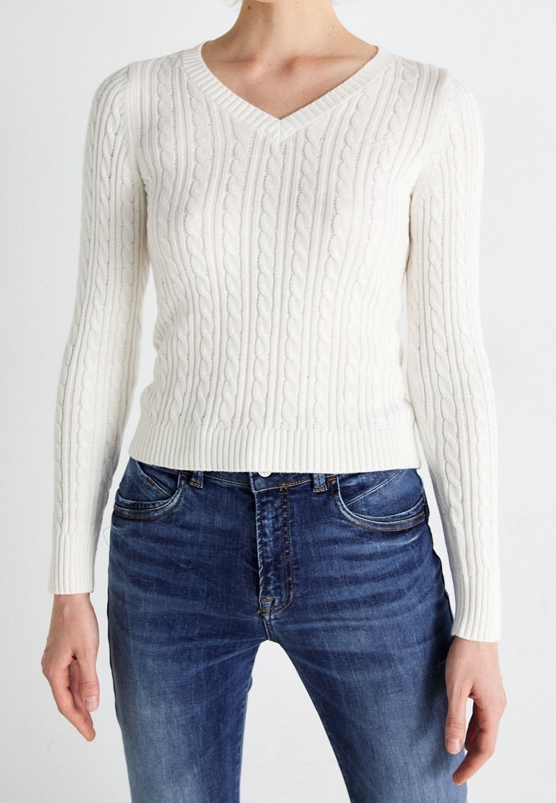 Femme portant un pull blanc ajusté en maille torsadée à col en V et un jean bleu, debout devant un fond blanc uni.