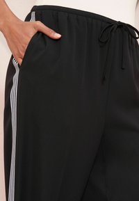 Čierne joggers nohavice s textúrovanou tkaninou, majú sťahovaciu pásku v páse, bočné vrecká a biele pruhy pozdĺž švov.