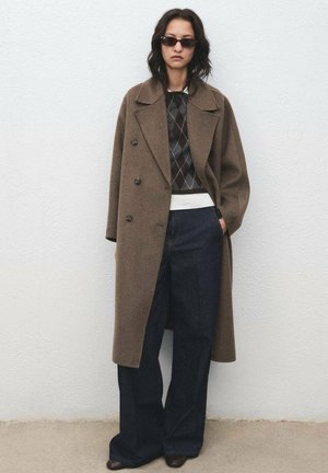 Cappotto doppiopetto in lana marrone, maglione a fantasia, vita bianca, pantaloni in denim scuri a gamba larga e occhiali da sole neri. Materiali testurizzati e vestibilità su misura.