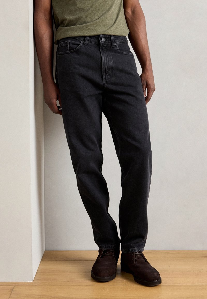Homme portant un jean noir droit, des chaussures marron foncé et un t-shirt vert olive, debout sur un sol en bois.