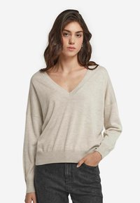 Beżowy sweter z długim rękawem i dekoltem w serek o luźnym kroju, z prążkowanymi mankietami i dół. Miękka tekstura, minimalistyczny design, w połączeniu z ciemnymi dżinsami.