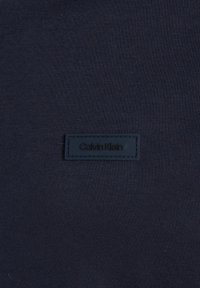 Marinfärgad tyg med en slät yta; har en sydd svart läderetikett med "Calvin Klein" pr präglat, placerad i nedre vänstra hörnet.