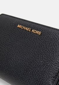 Juoda odinė piniginė su tekstūruota paviršiumi, priekyje esantis auksinis metalinis "MICHAEL KORS" logotipas. Kompaktiška ir stačiakampio formos.