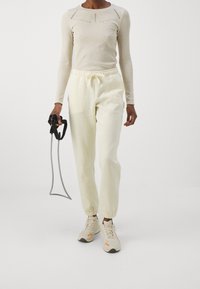 Person i beige långärmad tröja och krämfärgade joggers som håller i ett svart och grått motståndsband mot en vit bakgrund.