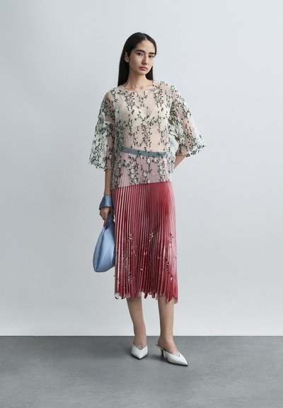 Haut transparent avec broderie florale, jupe plissée à plusieurs couches en dégradé de rouge, accessoirisée d'un sac bleu clair et de mules blanches à talons.