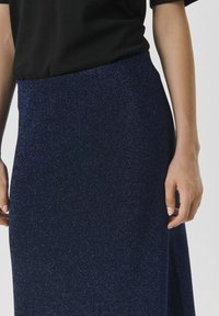 Fustă bleu marin cu o textură subtilă de sclipici, având un design drept și o fanta laterală. Asortată cu un top negru cu mânecă scurtă.