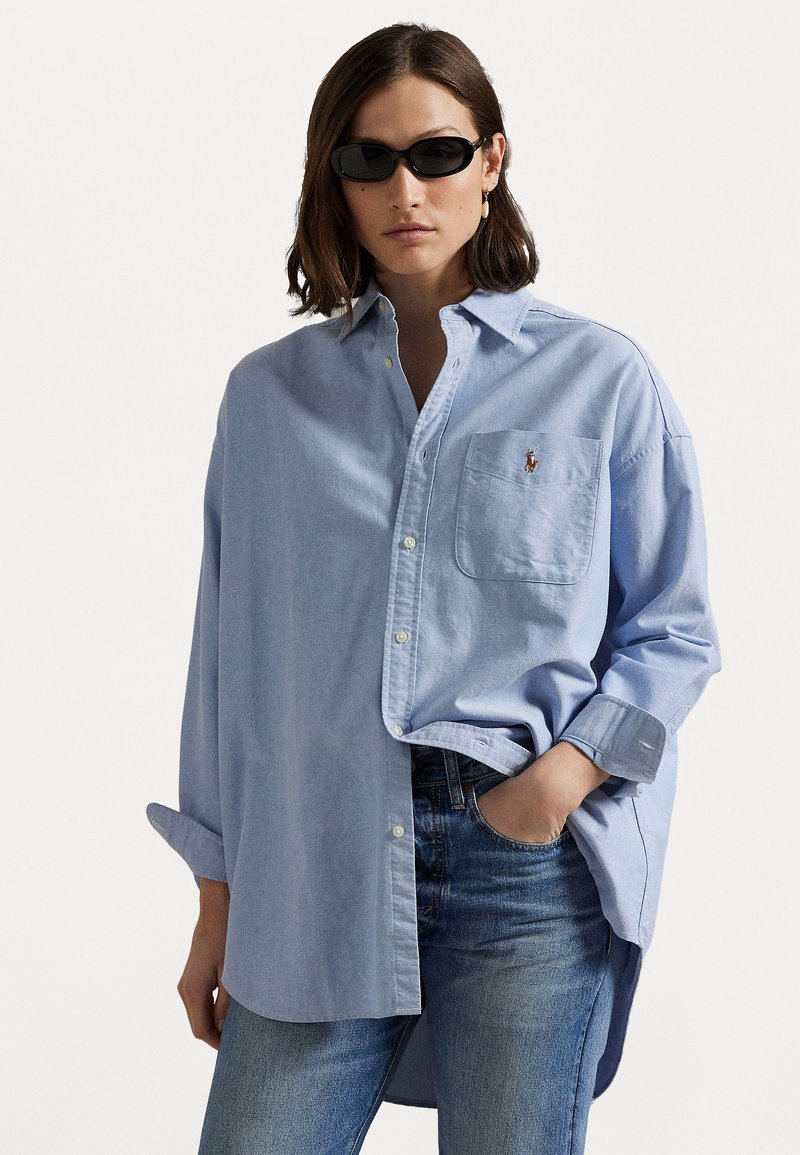 Lyseblå oversized knapp-up skjorte med brystlomme og brodert logo; kombinert med mørke jeans og svarte solbriller.