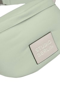 Fanny pack in tessuto verde chiaro con una texture liscia, caratterizzato da una patch rettangolare rosa con testo in rilievo. Incluso cinturino regolabile.