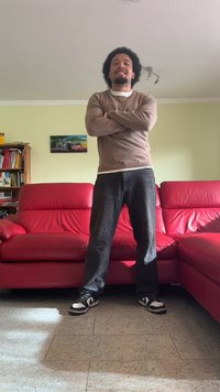 Une personne portant un pull marron, un jean noir et des baskets noir et blanc se tient dans une pièce avec des canapés en cuir rouge et un mur vert clair.