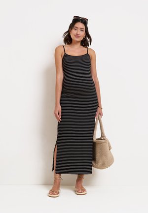 Femme portant une robe longue sans manches rayée noir et blanc avec une fente latérale, des sandales marron clair, des lunettes de soleil sur la tête et tenant un sac tissé beige.